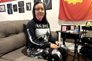 June Voros is sitting on a couch in her home. Her hands are placed on her knees and she looks towards the camera.