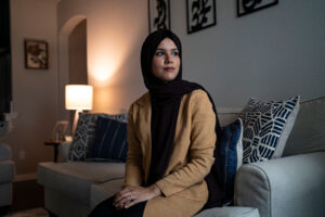 A woman sits on a couch with her hands folded in her lap and looks off-camera to her left.