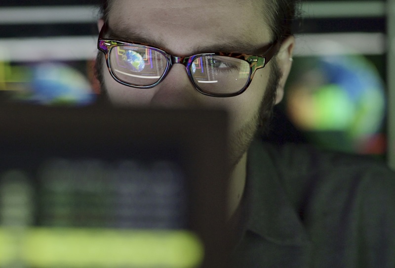 A bearded young man studying a computer screen displaying images of the earth with moving graphics. He’s surrounded by out of focus, colourful displays and reflections showing moving global graphics that are out of focus.