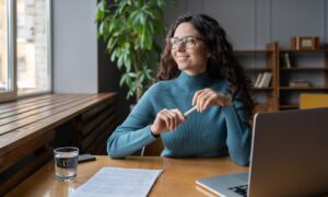 a woman smiling as she looks out a window. she has a laptop and a glass of water in front of her, alongside some paperwork