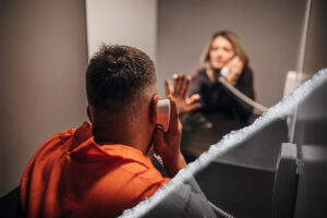 A woman talks to a prisoner; both have their hand on the glass divider between them.