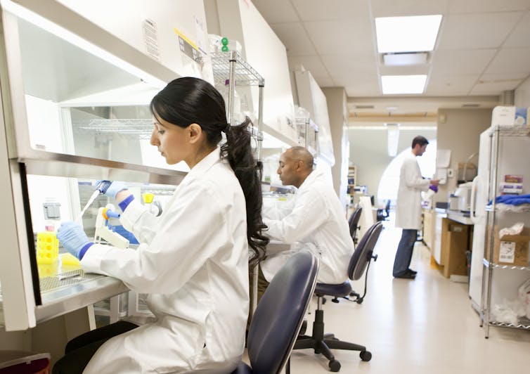 Two scientists pipetting liquids under a fume hood, with another scientist in the background examining a sample