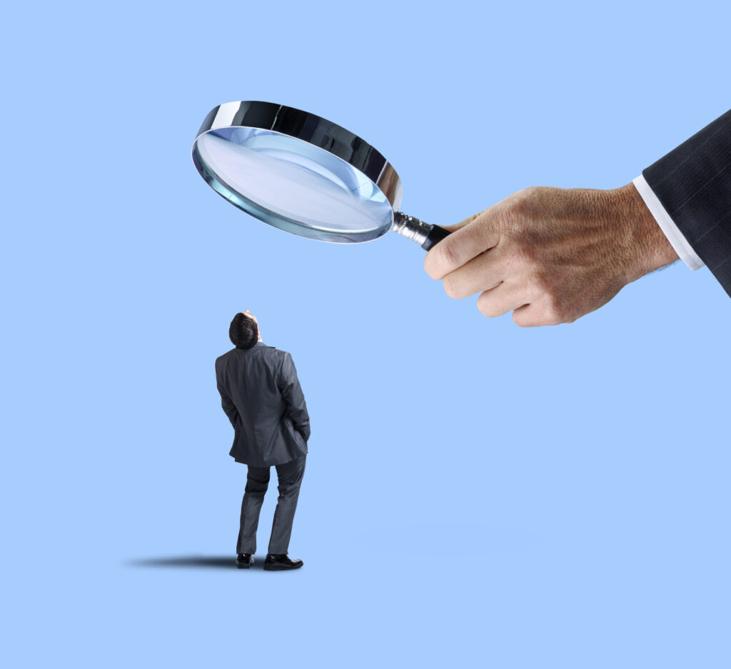 A businessman, under supervision, looks up at a large magnifying glass that is being held by large hand above him.