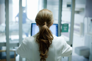 A photo of a woman seen from behind working in a medical setting on a laptop.