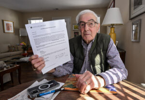 A senior man holds a letter from a Medicare provider. He is seated a table wearing glasses and a shirt and vest