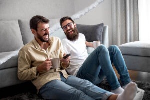 Two young men sit on the floor in front of a couch, just laughing and laughing as they hold coffee cups.