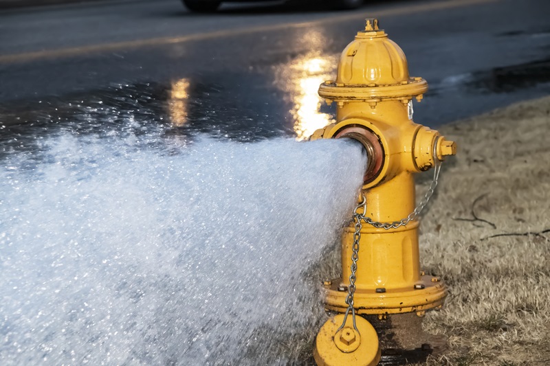 Close-up of yellow fire hydrant gushing water across a street with wet highway and tire from passing car behind