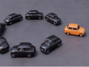 Aerial view of eight mini auto cars on asphalt: seven black cars driving in one direction and one orange car driving away from the pack