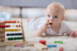 Baby chews on toy block while playing on the floor in her home. Toys and an abacus are blurred in the foreground.