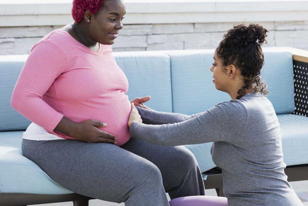 A pregnant African-American woman with her doula or birth support coach. The expectant mother, who had pink hair and is wearing a pink shirt, is sitting outdoors on a patio sofa smiling. The doula is kneeling in front of her, touching her round abdomen. Both women are in their 30s.