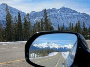 Side mirror of a moving car offers a picturesque view of the famous Icefields Parkway route and the stunning snowy mountains of Alberta. Car drives along scenic mountain road in Canada.