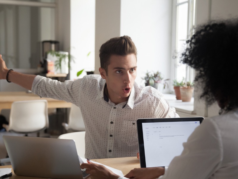 Frustrated customer expressing anger with arm outstretched in an expressive gesture during a conversation with a broker businessperson who is working on a laptop.