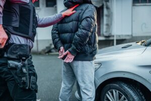 Police officer arresting a young gangster by the car in rainy weather.