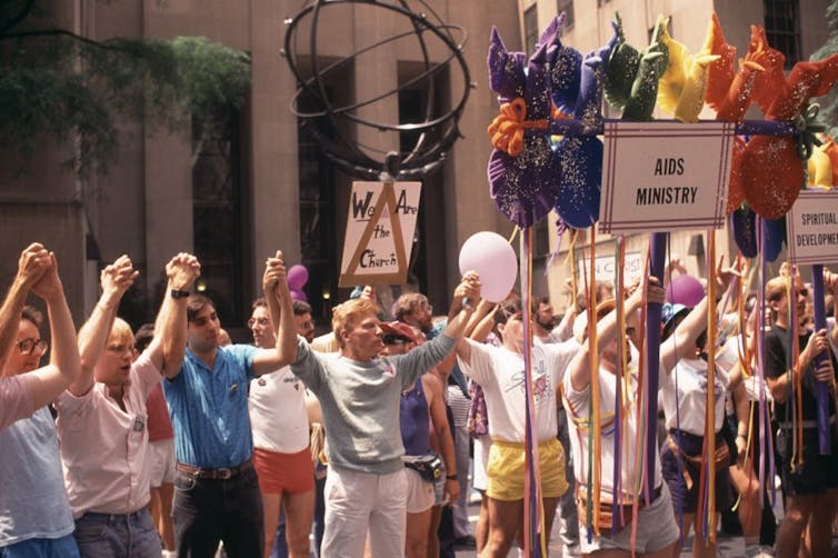 A group of men in colorful shirts stand in a row, raising their clasped hands, in front of a white building.