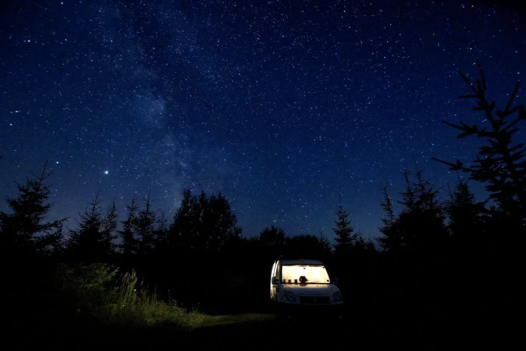 Van parked in wooded area at night, its lights glowing in the darkness