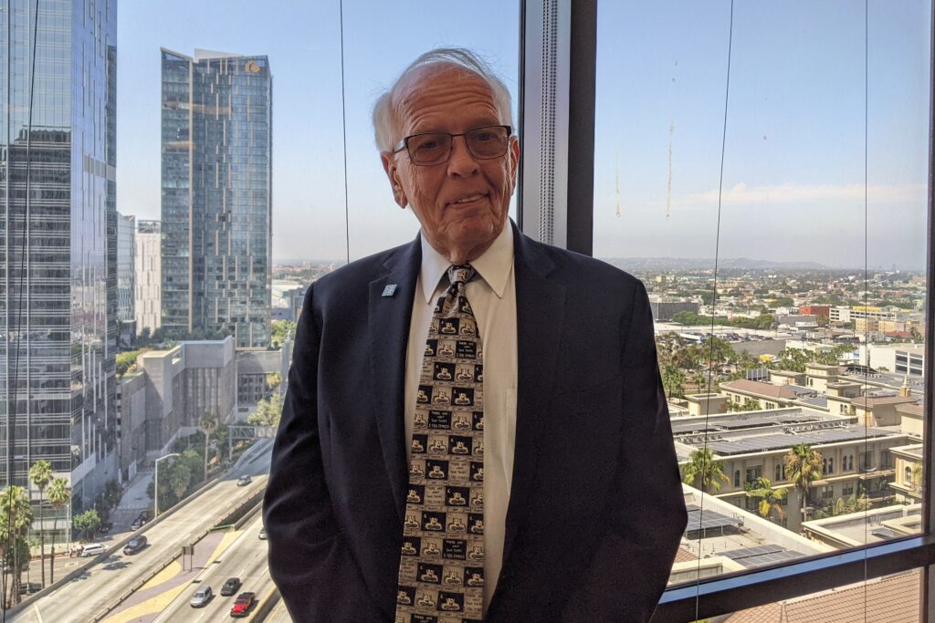 An older man in a suit stands in front of a large window with a view of skyscraper buildings