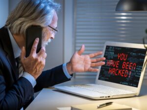 Back angle of a man with gray hair, glasses and a suit on the phone and gesturing in front of his laptop screen that reads "you've been hacked"