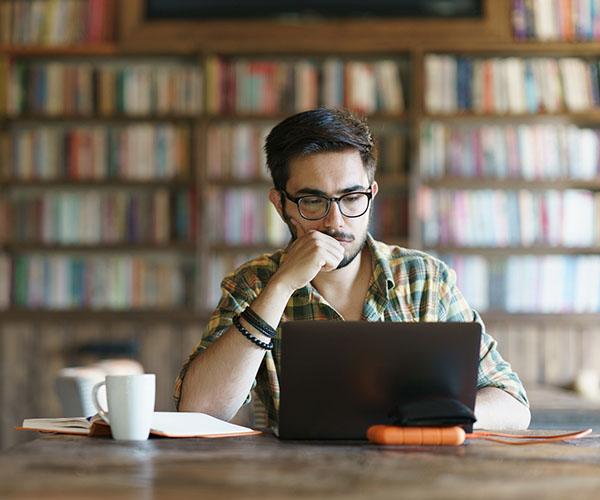 A person sitting at a table looking at a laptop with bookshelves behind them.