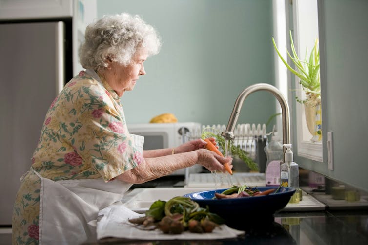 An elderly woman washes vegetables in the kitchen sink.