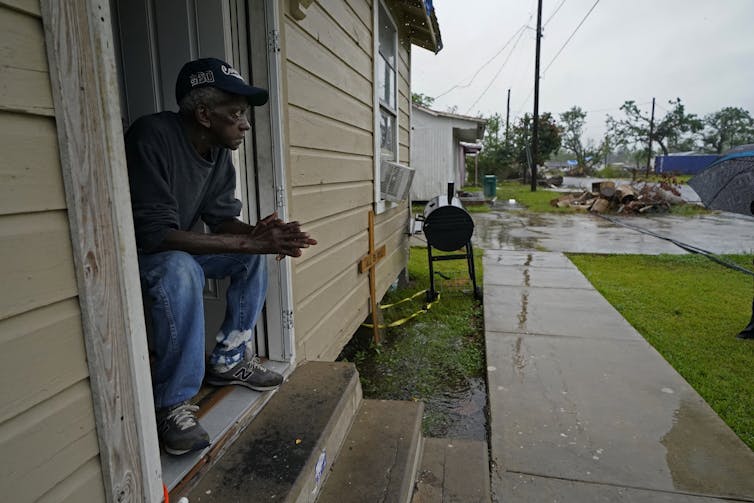 A man sits in the door of a house with a worried look on his face. Rain from Hurricane Delta is already falling.